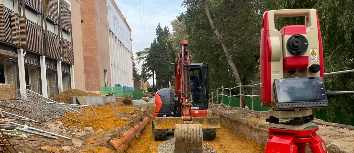 Estación total sobre trípode en una obra urbana, con una miniexcavadora trabajando en la zanja